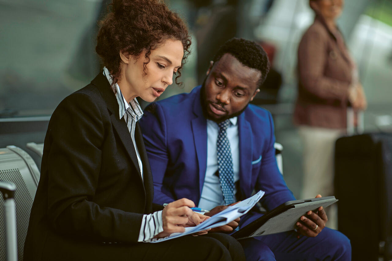 A man and a woman sit at the airport, reviewing notes on papers and tablets in preparation for a meeting